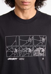 T-shirt en coton noir avec un graphisme blanc représentant des toiles d'araignée disposées en panneaux, avec le texte "CARHARTT WIP THEORY & PRACTICE" en bas.