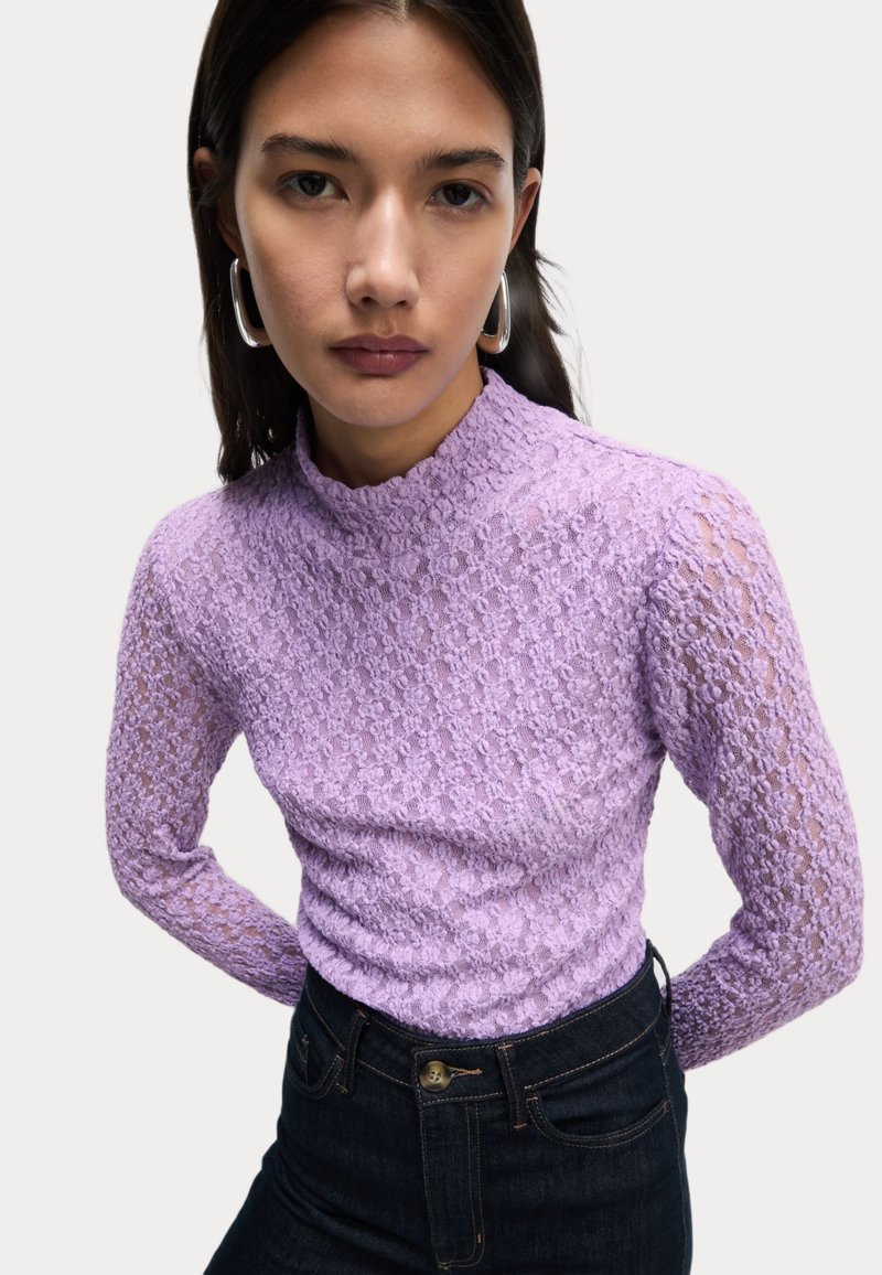Top lungo a maniche con pizzo viola, collo alto e motivo testurizzato, abbinato a jeans scuri in denim. Orecchini a cerchio argento aggiungono un tocco di dettaglio.