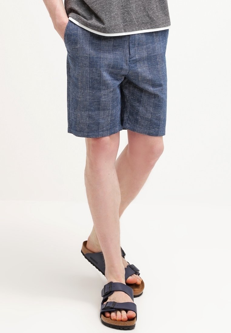 Shorts bleus pour hommes avec un motif à carreaux, en tissu léger. Associés à des sandales foncées avec des boucles ajustables.