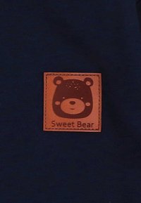 Fascia in pelle marrone con grafica di un orso e testo "Sweet Bear" su uno sfondo di tessuto blu scuro. Forma quadrata con bordi cuciti.