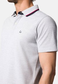 Jasnoszara koszulka polo z fakturowanej tkaniny, kontrastującym granatowym kołnierzykiem z czerwonymi akcentami i małym logo po lewej stronie klatki piersiowej.