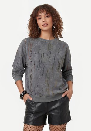 Kvinde med krøllet hår iført en grå slidt sweatshirt, sorte lædershorts, netstrømper og nittearmbånd, hænderne i lommerne.