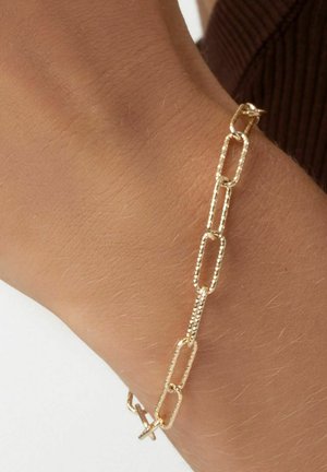 Guldkæde armbånd med forlægte, teksturerede led. Lukkes med en simpel lås og hviler på et håndled mod en lys baggrund.