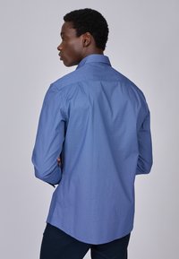 Chemise à manches longues à motif bleu avec col pointu et ourlet arrondi. Le tissu a une finition texturée avec un subtil design géométrique.