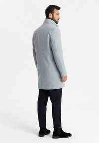 Abrigo de lana gris con cuello alto, diseño de un solo botón y silueta recta. Combinado con botas de tobillo negras y pantalones oscuros.