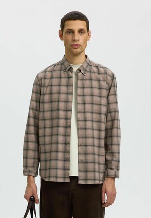 Homme portant une chemise à carreaux beige et grise boutonnée par-dessus une chemise blanche, tenant une sangle sombre dans une main, debout devant un fond uni.
