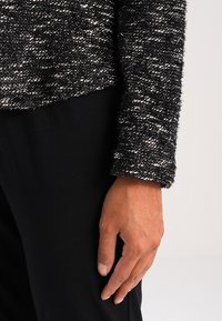 Pull texturé noir et blanc avec une finition duveteuse et une coupe légèrement oversize, présentant une longueur de manche décontractée, associé à un pantalon noir.