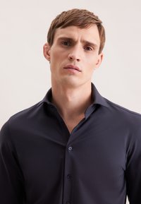 Camicia button-up blu scuro con una texture elegante e liscia, presentata con colletto e bottoni, indossata da un modello su uno sfondo neutro.
