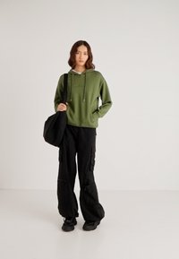 Zelená mikina s dlhými rukávmi a sťahovacou šnúrkou, v kombinácii s čiernymi cargo nohavicami s bočnými vreckami. Čierna taška na rameno a tenisky.