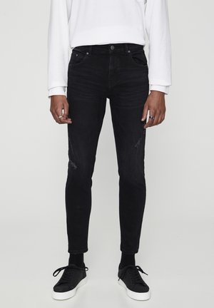 Personne portant un jean noir slim-fit légèrement usé, une chemise blanche à manches longues, des chaussettes noires et des baskets noires avec des semelles blanches.