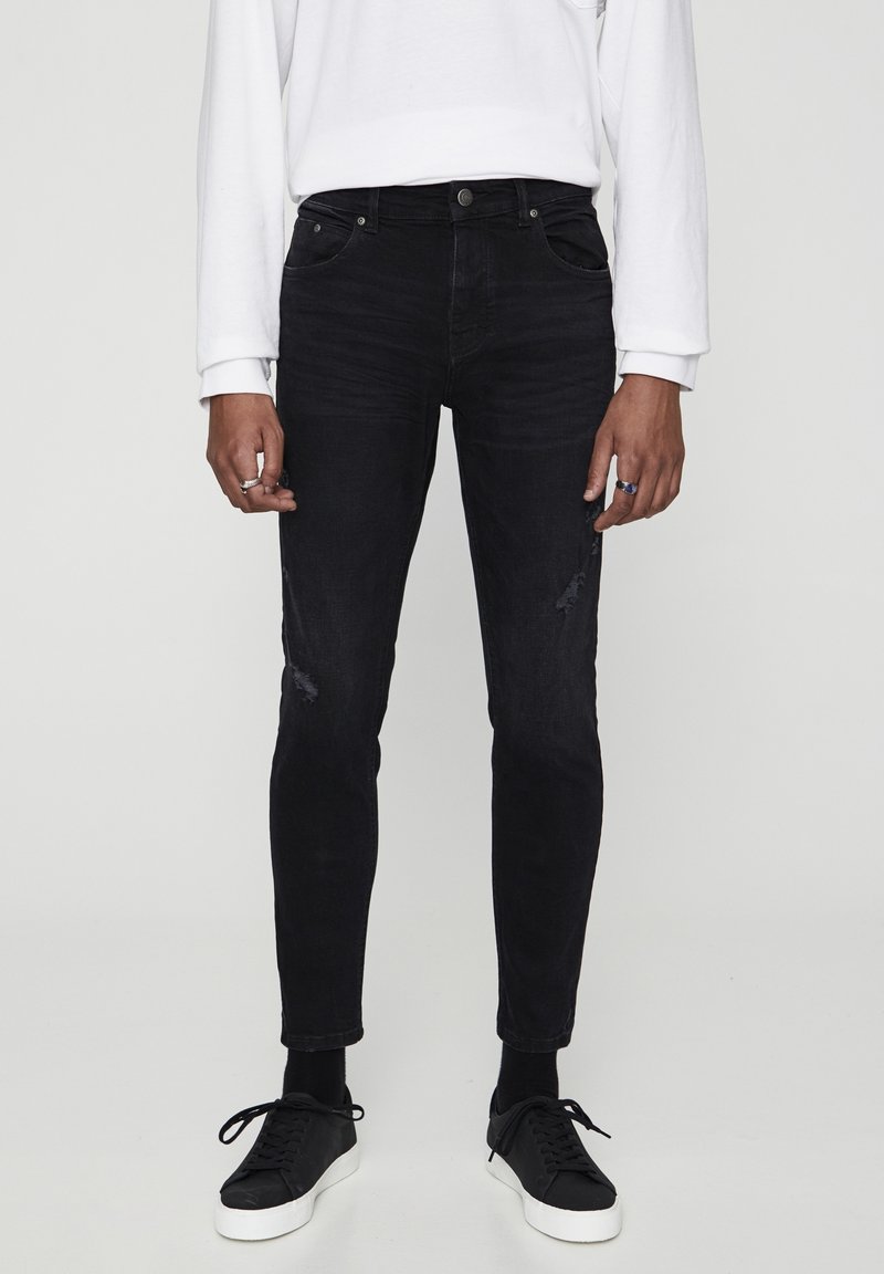 Personne portant un jean noir slim-fit légèrement usé, une chemise blanche à manches longues, des chaussettes noires et des baskets noires avec des semelles blanches.