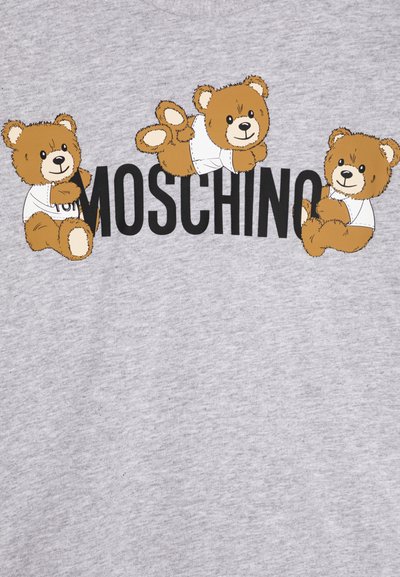 Szürke póló, amelyen három mesefigurás medve látható fehér ingben, a fekete "MOSCHINO" felirat felett. Puha pamut anyag.