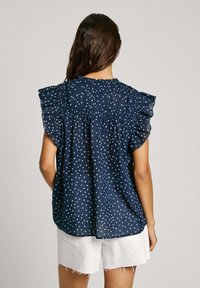 Námornícky modrá blúzka s bielymi bodkami, volánovými rukávmi a zberaným chrbtom, vyrobená z ľahkého materiálu. Stylizovaná s bielymi šortkami.