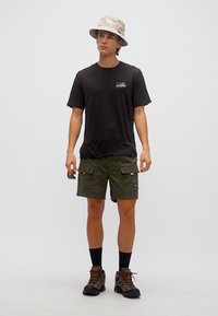 Νεαρός άνδρας που φοράει σκουρόχρωμη μπλούζα, χακί σορτς cargo, μπεζ καπέλο τύπου κάπα, μαύρες κάλτσες και παπούτσια πεζοπορίας, κρατώντας γυαλιά ηλίου στο ένα χέρι.