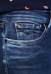 Jeans de denim azul oscuro con un bolsillo frontal, que presentan detalles de costura y un cierre de botón de cobre. Textura suave con un ligero desvanecimiento.