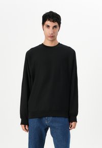 Felpa nera a maniche lunghe in un tessuto morbido con collo a giro. Presenta polsini elasticizzati e un logo discreto sul petto. Abbinata a jeans blu.
