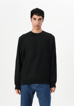 Svart langermet genser i et mykt stoff med rund hals. Har elastiske mansjetter og en diskret logo på brystet. Kombinert med blå jeans.