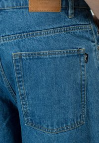 Los pantalones vaqueros presentan un color azul con una textura ligeramente desgastada, un bolsillo trasero y una etiqueta de cuero con un logotipo en relieve en la cinturilla.