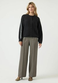 Svart stickad kofta med knäppning och vit dekorsöm på ärmarna, tillsammans med ljusgrå vida byxor.