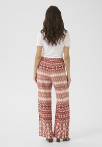 Pantalons évasés à motifs arborant un mélange de teintes rouge et crème avec des formes géométriques et des accents floraux. Portés avec un haut blanc.