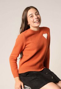 Pull orange avec un col rond et une appliqué en forme de cœur blanc sur la poitrine gauche. Poignets et ourlet côtelés, texture tricot douce.