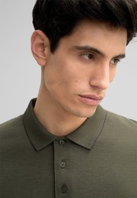 Dunkelgrünes Polohemd aus strukturiertem Stoff, mit klassischem Kragen, fünf Knöpfen und Kontrastnähten um den Kragen.