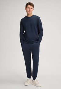 Navy blue long-sleeve sweatshirt with round neck and logo patch; paired with matching joggers; features elastic cuffs and waistband; white sneakers.