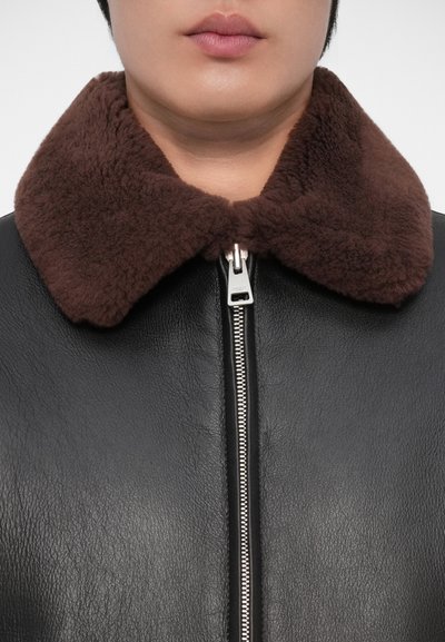 Zwarte leren jas met een bruin schapenvacht kraag en een zilveren ritsluiting. De textuur is zacht met een subtiel nerfpatroon.
