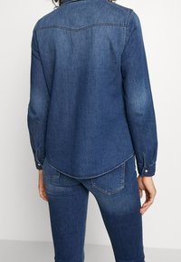 Mörkblå denimskjorta med långa ärmar, knappmanschetter, sicksackad fåll och yoke-detaljer på ryggen. Slät textur med en klassisk design.