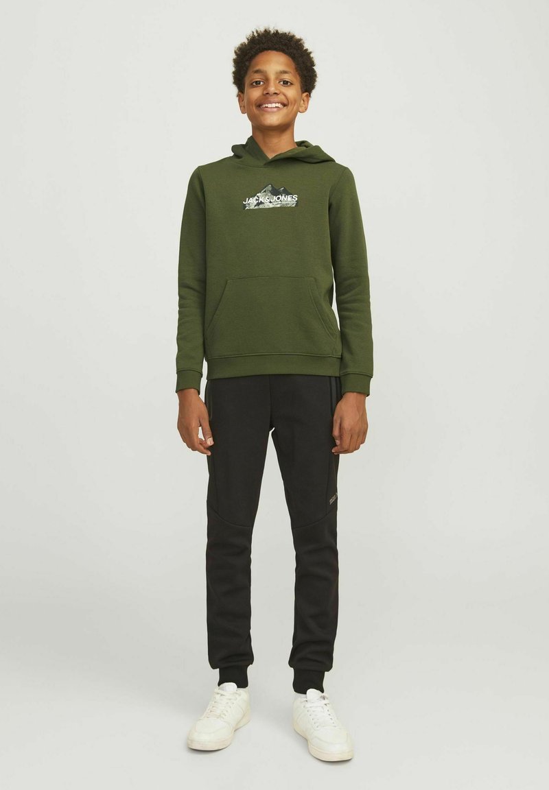 Felpa verde con cappuccio, dotata di tasca frontale e logo. Abbinata a pantaloni della tuta neri a gamba affusolata con cuciture decorative e sneakers bianche.