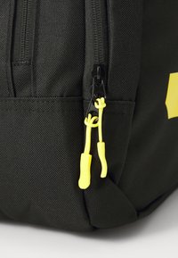 Black fabric backpack featuring a zipper with black hardware and yellow pull cords. Textured exterior with an angular design element.