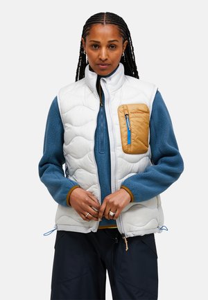 Gilet bianco isolato con texture trapuntata, dotato di una tasca sul petto color cammello e una cerniera blu, indossato sopra un maglione in pile blu. Pantaloni neri sotto.