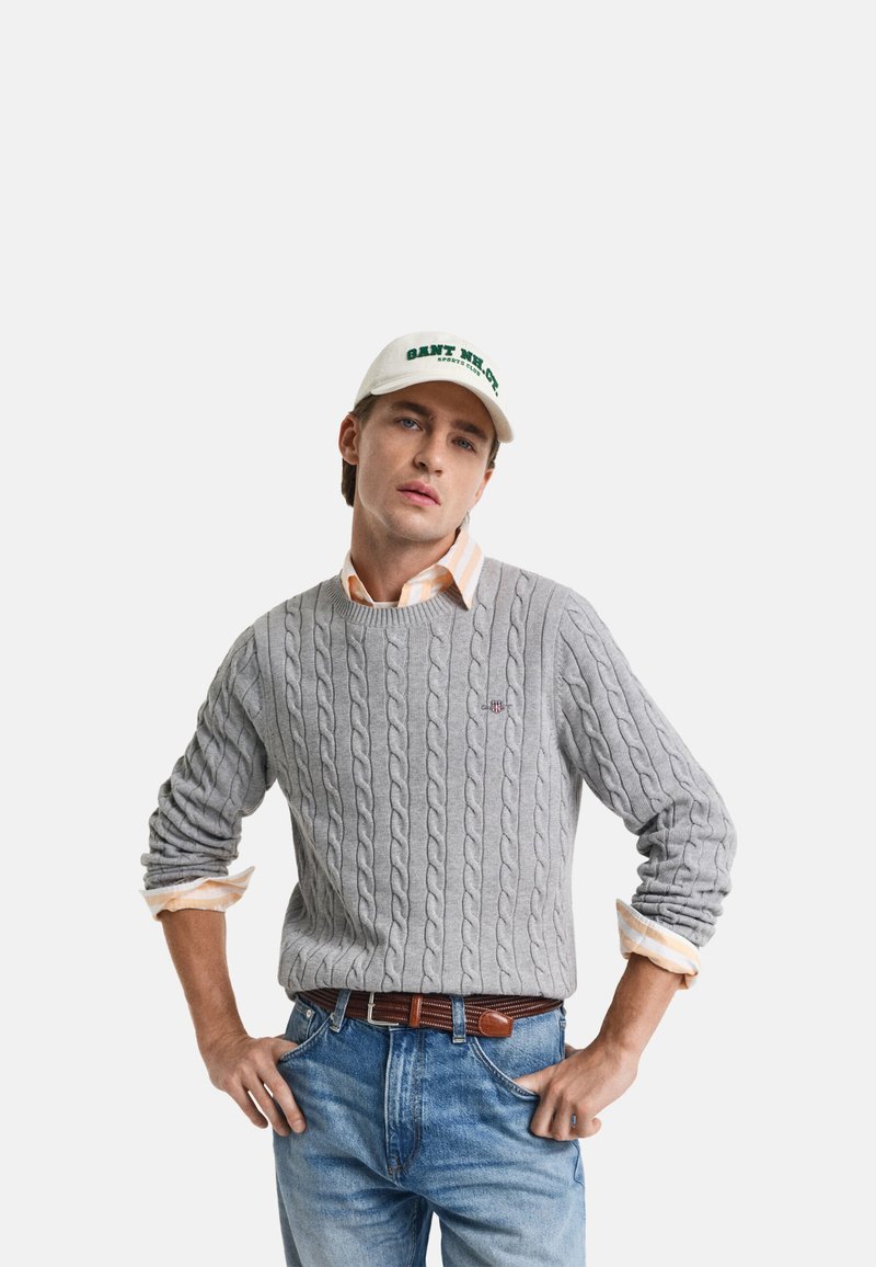 Grå kabelstrikket sweater med ribbede kant, lagd over en orange og hvid ternet skjorte. Kombineret med lyseblå jeans og en hvid kasket.