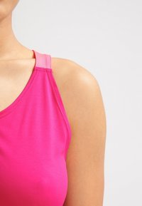 Ingrandimento di una persona che indossa una maglietta senza maniche di un vivace colore rosa, con le spalle e la parte superiore del torace visibili su uno sfondo chiaro e uniforme.