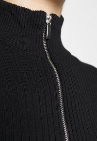 Svart ribbad tröja med hög krage, med detaljer av silvrig dragkedja. Tyget har en slät yta med vertikala ränder.