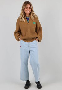 Sweat à zip marron côtelé avec patch logo vert, associé à un jean large bleu clair et des bottes noires. Fond neutre.
