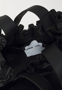 Interno di una borsa in pelle nera con bordi arricciati, caratterizzato da un'etichetta bianca con la scritta "SAMSNØ SAMS". Texture morbida e linee curve visibili.