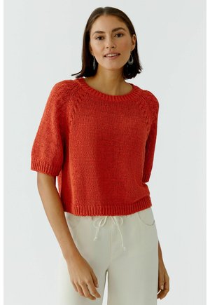 Frau trägt roten kurzärmeligen Strickpullover und weiße Hose mit Kordelzug, steht vor hellem, einfarbigem Hintergrund und blickt nach vorn.