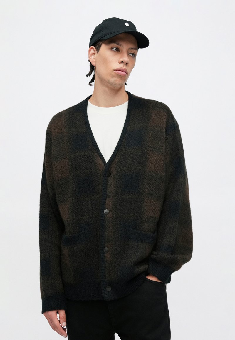 Dark brown and navy checkered cardigan, knitted texture, v-neck, featuring three buttons and two front pockets. Black cap worn above.