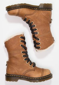 Botas de cuero marrón con cordones, bordeadas de piel sintética, cordones negros, ojales dorados, parte superior texturizada y una suela negra duradera con costuras naranjas.