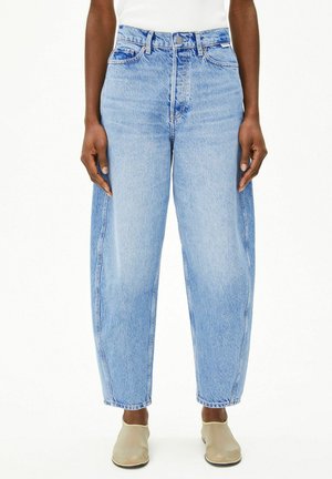 Hellblaue Jeans mit hoher Taille und geradem Bein, getragen mit beigen Slippern bei einer Person, die vor weißem Hintergrund steht.