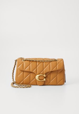 Kuldse ketiga kandmine tan värvi pehmest nahast käekott, mille esiküljel on silmapaistev kuldne ringikujuline lukk.