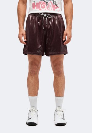 Mann trägt dunkle glänzende Basketballshorts mit weißem Kordelzug, weiße Socken und weiße Sportschuhe und steht vor einem schlichten Hintergrund.