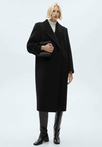 Cappotto di lana nera con un design strutturato, ampi rever e chiusura a doppio bottone. Abbinato a stivali lucidi neri al ginocchio.