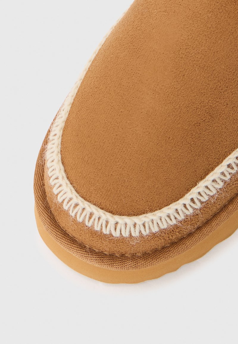 Zamszowe buty w kolorze tan, o zaokrąglonym kształcie, z białym haftem dookoła krawędzi oraz gumową podeszwą zapewniającą przyczepność.