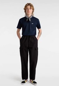 Polo azul marino con logo blanco, pantalones cargo negros con bolsillos, zapatillas negras y multicolores, bufanda de cuadros alrededor del cuello.