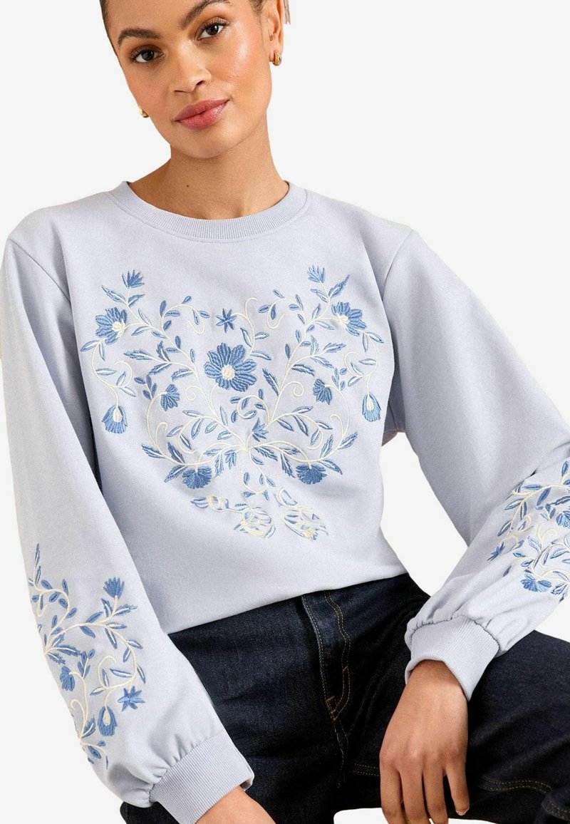 Svetlo modra majica s cvetnim vezenjem v temno modri in beli barvi na sprednji strani in rokavih. Rebrast ovratnik in elastični zaključki.