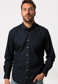 Merkkipaitana on puuvillainen tummansininen napitettava paita, jossa on pitkät hihat, etutasku ja valkoiset napit. Tekstuuriltaan sileä ja leikkaukseltaan istuva.