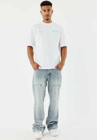 Camiseta blanca de gran tamaño con gráficos en azul claro, combinada con jeans baggy azul desvaído y zapatillas blancas. Diseño simple y ajuste casual.