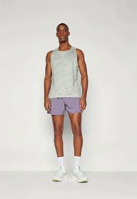 Šviesiai pilkas tank topas su bangų raštu, derinamas su levandų spalvos šortais. Modelis dėvi baltus sportinius batelius ir baltas kojines, stovintis prieš neutralų foną.